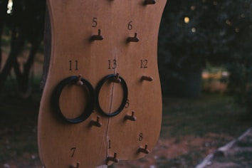 A wooden ring toss board with pegs numbered 1 to 13, two black rings hanging on the pegs labeled 11 and 13. The background features a blurred outdoor scene with dark foliage.