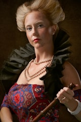 A person with an elegant hairstyle, wearing a richly decorated dress with red and blue patterns and adorned with pearl necklaces. The individual is holding a wooden cane or staff and has a confident expression. The ruffled black collar adds to the dramatic effect.