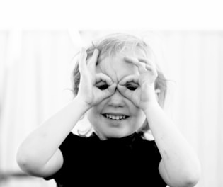 A young child with light hair makes a playful gesture by forming circles with their fingers around their eyes, resembling binoculars. The child is smiling and appears to be enjoying themselves. The image is black and white, adding a classic touch.