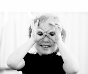 A young child with light hair makes a playful gesture by forming circles with their fingers around their eyes, resembling binoculars. The child is smiling and appears to be enjoying themselves. The image is black and white, adding a classic touch.