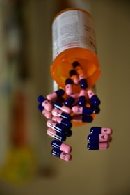 An orange prescription pill bottle lies on its side with several blue and pink capsules scattered around it. The capsules are marked with the label 'A 4'. The bottle is plastic and white with a readable label, suggesting a medical or pharmaceutical context.