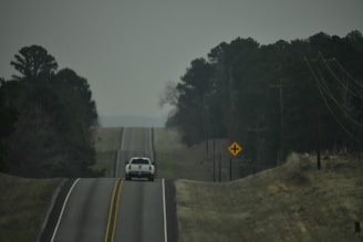 A white pickup truck is driving down a rural, two-lane road. The road appears hilly with slight elevation changes, and it is flanked by trees and power lines. The sky is overcast, and a road sign indicating an upcoming intersection is visible on the right side.