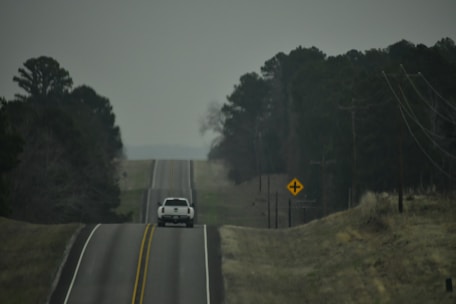 A white pickup truck is driving down a rural, two-lane road. The road appears hilly with slight elevation changes, and it is flanked by trees and power lines. The sky is overcast, and a road sign indicating an upcoming intersection is visible on the right side.