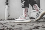 A close-up of a person wearing sporty, patterned sneakers with 'Supply 350' branding and white socks featuring the word 'Skepticism' in bold red letters. The setting is an outdoor paved area next to a light-colored wall with some scattered dry leaves on the ground.