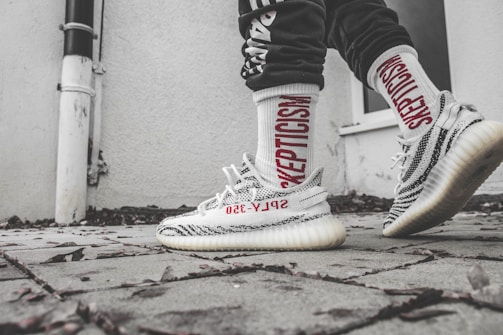 A close-up of a person wearing sporty, patterned sneakers with 'Supply 350' branding and white socks featuring the word 'Skepticism' in bold red letters. The setting is an outdoor paved area next to a light-colored wall with some scattered dry leaves on the ground.