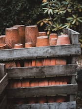 Several clay flower pots are stacked upside down inside a weathered wooden crate, surrounded by greenery with blurred foliage in the background. The pots have an aged and rustic appearance.