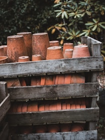Several clay flower pots are stacked upside down inside a weathered wooden crate, surrounded by greenery with blurred foliage in the background. The pots have an aged and rustic appearance.