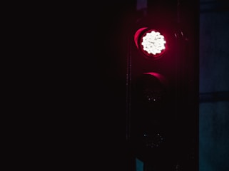 A traffic light displays a bright red light against a dark background, indicating a stop signal. The surroundings are dimly lit, emphasizing the glow of the red light.