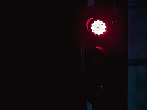 A traffic light displays a bright red light against a dark background, indicating a stop signal. The surroundings are dimly lit, emphasizing the glow of the red light.