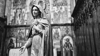 A black and white image of a religious statue depicting a robed figure with a halo, standing in an ornate church interior. The background features detailed religious icons and carved wooden structures.