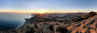 A panoramic view captures a coastal cityscape at sunset. The image features a rocky foreground leading into a sprawling urban area bordered by the sea. The horizon shows mountains under a clear sky, with the sun setting and casting warm hues across the landscape.