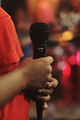 A person holds a black microphone in their hand. The background features blurred lights in various colors, primarily red and orange. The person is wearing a red shirt, and the lighting suggests a warm, indoor setting, possibly during a performance or event.