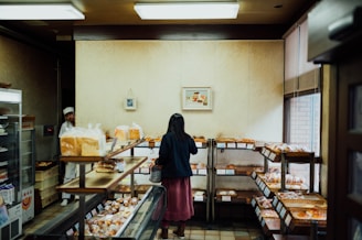 A woman is browsing in a cozy bakery, examining various baked goods displayed on wooden shelves. The bakery has a warm, inviting atmosphere with soft lighting. A man in a white apron and hat stands near the entrance, possibly a staff member.
