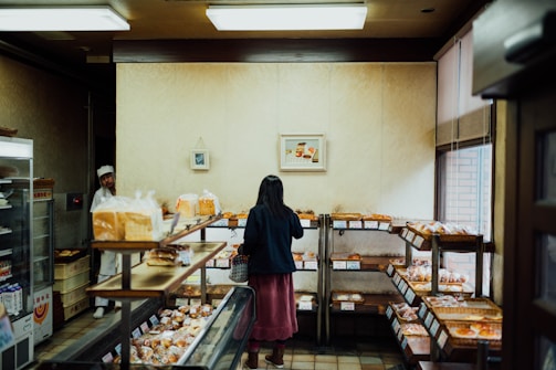 A woman is browsing in a cozy bakery, examining various baked goods displayed on wooden shelves. The bakery has a warm, inviting atmosphere with soft lighting. A man in a white apron and hat stands near the entrance, possibly a staff member.