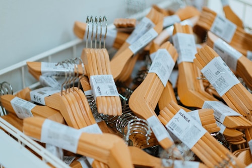 A large pile of wooden coat hangers with metal hooks is seen inside a white storage bin. Each hanger has a white label wrapped around it containing some text and barcodes.