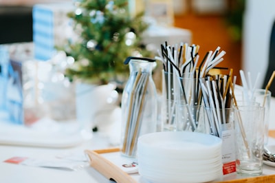A table with various party supplies is neatly arranged, featuring a small Christmas tree in the background. Several glasses and a jug are filled with black, white, and gold drinking straws. A stack of white plates is placed on a wooden tray nearby.