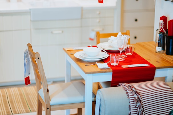 A neatly set dining table with a wooden surface is adorned with a red table runner. The table contains white plates, bowls, and drinking glasses. A bottle and red candles are placed at the side. Two wooden chairs with white cushions are visible, and a striped blanket hangs over one chair.
