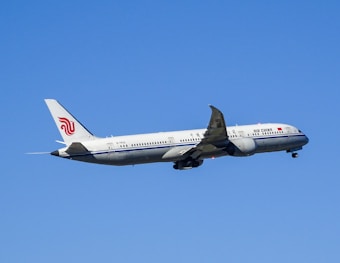 An airplane with Air China livery is flying against a clear blue sky. The aircraft's wings and tail are visible, with engines mounted under each wing. The plane is in ascent as indicated by its upward angle.