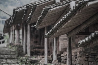A series of wooden structures with traditional tiled roofs supported by wooden beams. The scene includes a stone pathway with grass and plants poking through. The overall setting appears to be rustic and old, possibly in a rural or historical area.