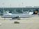 A Lufthansa airplane is parked on the tarmac in front of an airport terminal. The terminal building features large glass windows. Several light poles are visible, with a taxi positioned in front of the airplane.