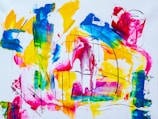 Abstract painting with vibrant colors including yellow, blue, pink, and green. The composition includes broad, sweeping brushstrokes and splashes of paint, intersected by dynamic black lines. The elements blend and interact, creating a visually engaging and energetic scene.