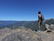 A person stands on a rocky cliff overlooking a vast, mountainous landscape with a bright blue sky. The individual is wearing hiking gear, including a backpack, and gazes towards the distant mountains, emphasizing a sense of adventure and exploration.