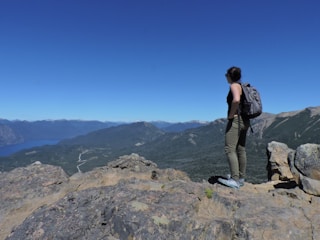A person stands on a rocky cliff overlooking a vast, mountainous landscape with a bright blue sky. The individual is wearing hiking gear, including a backpack, and gazes towards the distant mountains, emphasizing a sense of adventure and exploration.