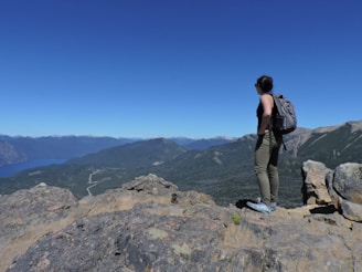 A person stands on a rocky cliff overlooking a vast, mountainous landscape with a bright blue sky. The individual is wearing hiking gear, including a backpack, and gazes towards the distant mountains, emphasizing a sense of adventure and exploration.