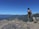 A person stands on a rocky cliff overlooking a vast, mountainous landscape with a bright blue sky. The individual is wearing hiking gear, including a backpack, and gazes towards the distant mountains, emphasizing a sense of adventure and exploration.