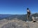 A person stands on a rocky cliff overlooking a vast, mountainous landscape with a bright blue sky. The individual is wearing hiking gear, including a backpack, and gazes towards the distant mountains, emphasizing a sense of adventure and exploration.