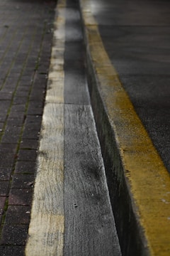 A wet roadway and sidewalk with a yellow line marking the edge of the road. The pavement appears damp, reflecting dim ambient light. The surface is textured with visible wear and small debris on the path.