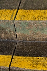 A cracked, weathered concrete surface with moss and two bright yellow lines painted across it. The crack runs vertically through the middle of the image.