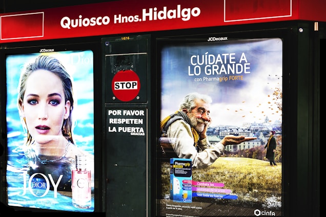There are two adjoining advertisements at a kiosk. On the left is an advertisement featuring a person with light hair in front of an aquatic background, promoting a perfume called 'JOY by Dior.' On the right is an advertisement depicting an elderly man with a warm smile holding a wooden bowl against a backdrop of a field with autumn leaves, promoting Pharmagrip Forte.