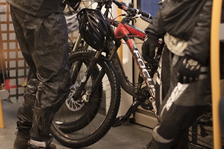 Two individuals dressed in dark, waterproof cycling gear are standing next to a mountain bike with a red and black frame. The setting appears to be indoors, possibly on a train or tram, indicated by the metal grating and seating. The bike is positioned vertically with a helmet resting on the handlebars.