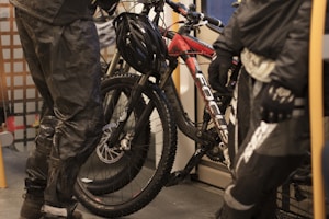 Two individuals dressed in dark, waterproof cycling gear are standing next to a mountain bike with a red and black frame. The setting appears to be indoors, possibly on a train or tram, indicated by the metal grating and seating. The bike is positioned vertically with a helmet resting on the handlebars.