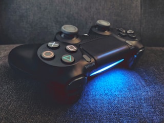 A black video game controller rests on a dark surface, illuminated from below by a vibrant blue light. The controller features recognizable buttons and directional pads, indicating it's designed for gaming consoles.