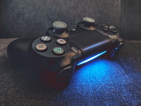 A black video game controller rests on a dark surface, illuminated from below by a vibrant blue light. The controller features recognizable buttons and directional pads, indicating it's designed for gaming consoles.