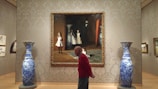 A large, ornately-framed painting depicts three young girls in white dresses, one seated and two standing, positioned next to a large ceramic vase. The painting is centered on a patterned wall flanked by two tall, blue and white porcelain vases. A person in a red sweater walks in front of the painting, their profile partially visible.