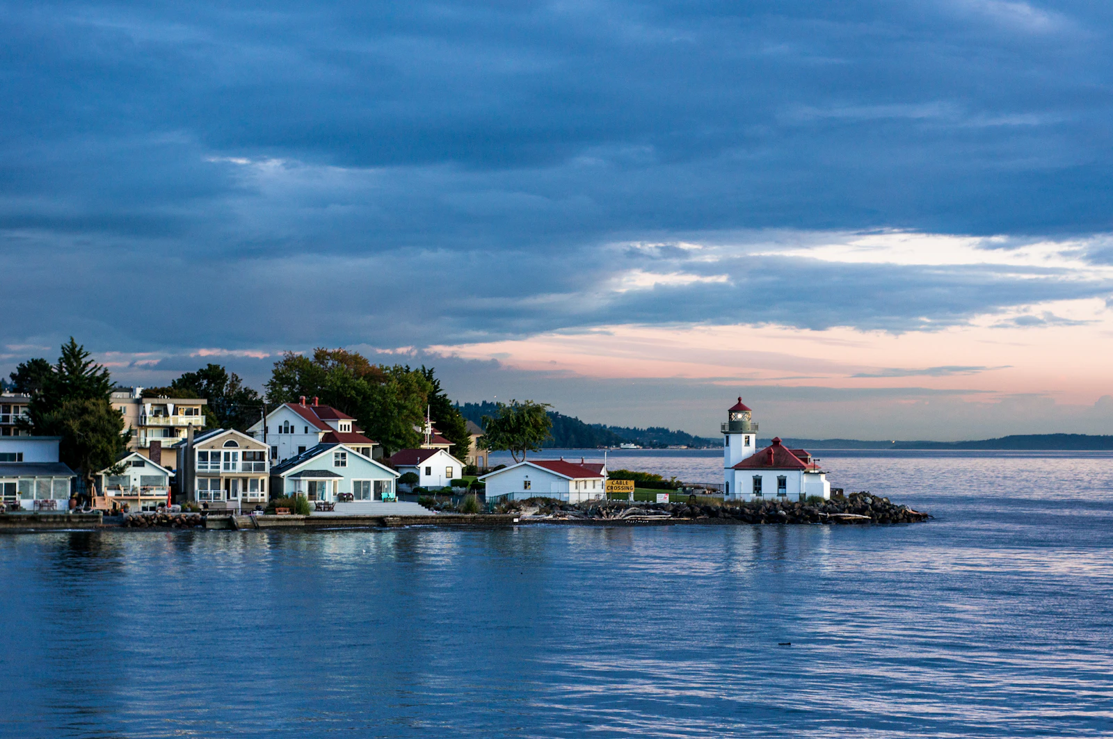 White lighthouse and clustered waterfront homes on a calm point at dusk, like Alki Point in West Seattle