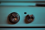Two metallic control knobs on a blue-green colored appliance or machine panel, with a dark shadow over one side. The larger knob is marked with 'OFF' and 'OPEN', while the smaller knob also has indicators. A small black dot is located between the two knobs.