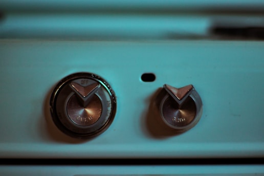 Two metallic control knobs on a blue-green colored appliance or machine panel, with a dark shadow over one side. The larger knob is marked with 'OFF' and 'OPEN', while the smaller knob also has indicators. A small black dot is located between the two knobs.