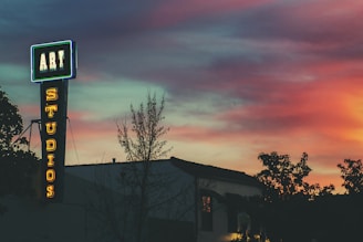 A neon sign that reads 'ART STUDIOS' stands out against a vibrant sunset sky. The sign is affixed to the side of a building and is surrounded by silhouettes of trees. The sky displays a gradient of colors, ranging from deep blue to warm hues of pink and orange.