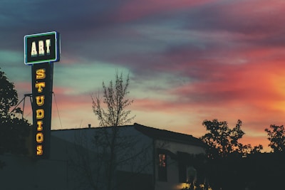 A neon sign that reads 'ART STUDIOS' stands out against a vibrant sunset sky. The sign is affixed to the side of a building and is surrounded by silhouettes of trees. The sky displays a gradient of colors, ranging from deep blue to warm hues of pink and orange.
