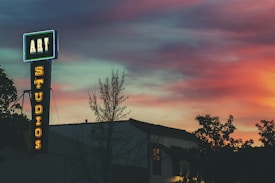 A neon sign that reads 'ART STUDIOS' stands out against a vibrant sunset sky. The sign is affixed to the side of a building and is surrounded by silhouettes of trees. The sky displays a gradient of colors, ranging from deep blue to warm hues of pink and orange.