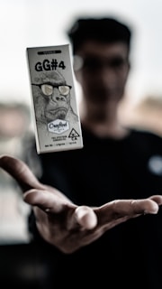 A hand is outstretched with a small box hovering above it. The box features an image of a gorilla wearing glasses and contains text such as 'GG#4' and 'Crafted Extracts'. The background is blurred, suggesting movement or focus on the foreground object.