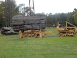 A large piece of timber is secured on a vintage yellow caterpillar tractor in a grassy field. The surrounding area features tall trees, a fence, and a rural landscape.