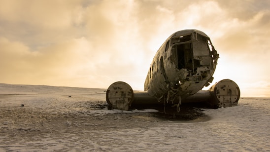 A wrecked airplane sits abandoned on a desolate, snow-covered landscape under an expansive, cloudy sky. The aircraft's fuselage is heavily weathered, with visible signs of rust and decay. The scene is lit by a soft, diffused light from the overcast sky, creating a somber and eerie atmosphere.