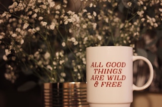 A white mug with red text saying 'ALL GOOD THINGS ARE WILD & FREE' is placed in front of a blurred background of delicate white flowers and greenery inside a metallic vase.