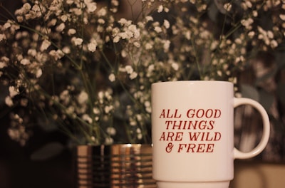 A white mug with red text saying 'ALL GOOD THINGS ARE WILD & FREE' is placed in front of a blurred background of delicate white flowers and greenery inside a metallic vase.