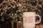 A white mug with red text saying 'ALL GOOD THINGS ARE WILD & FREE' is placed in front of a blurred background of delicate white flowers and greenery inside a metallic vase.